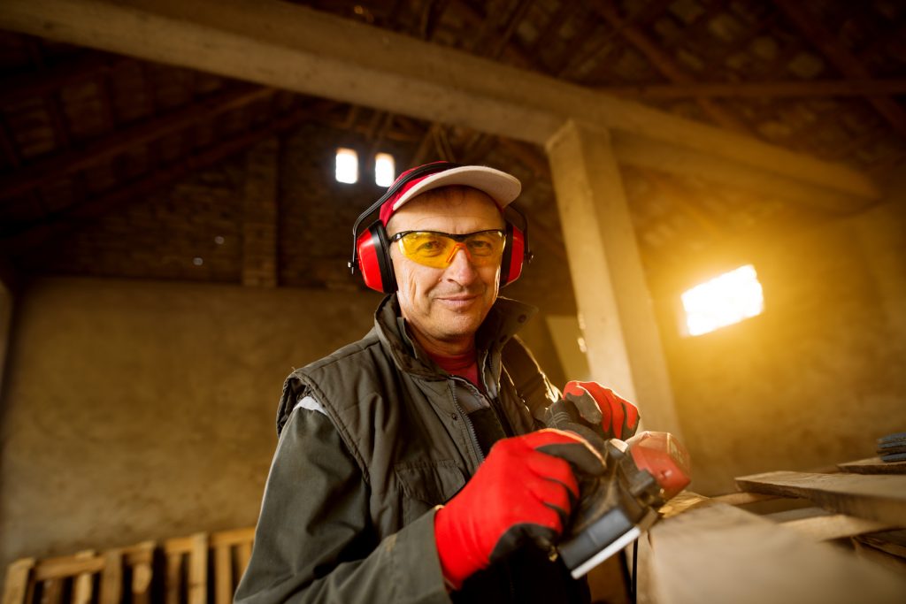 Modern senior carpenter in a professional uniform and protection working with an electric tool on a wood pallet. Schreiner